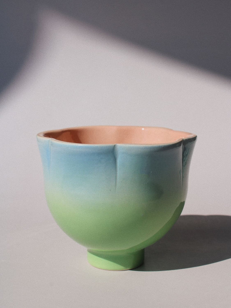 A ceramic cup with a scalloped rim and a small pedestal base, featuring a smooth gradient glaze transitioning from light blue at the top to soft green at the bottom, with a peach-colored interior, set against a neutral gray background with artistic shadows.