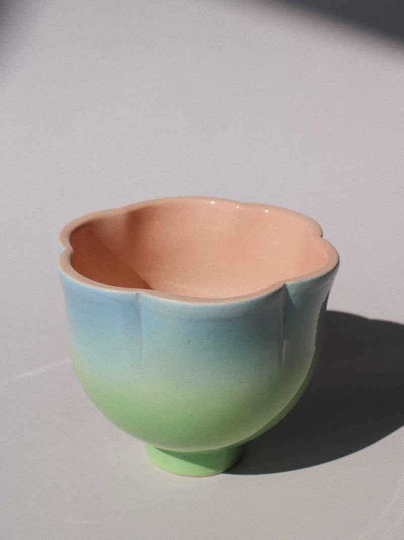 A ceramic flower-shaped cup with a smooth gradient glaze transitioning from peach-pink at the top to light blue and mint green at the bottom, set against a plain light gray background with a soft shadow.
