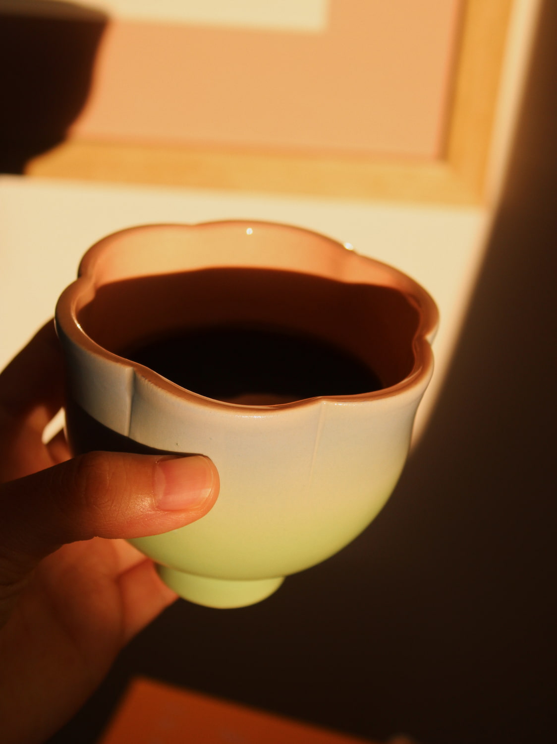 A hand holding a ceramic tea cup with a scalloped rim, featuring a light green to white gradient exterior and a warm pinkish-brown interior, lit by warm directional sunlight.
