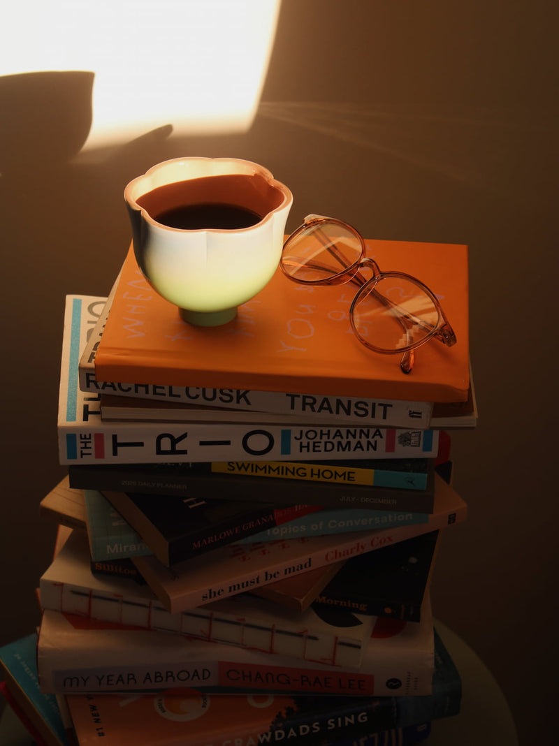 A stack of books on a table with a glowing ceramic cup and a pair of eyeglasses resting on top, lit by a warm spotlight against a dark background.