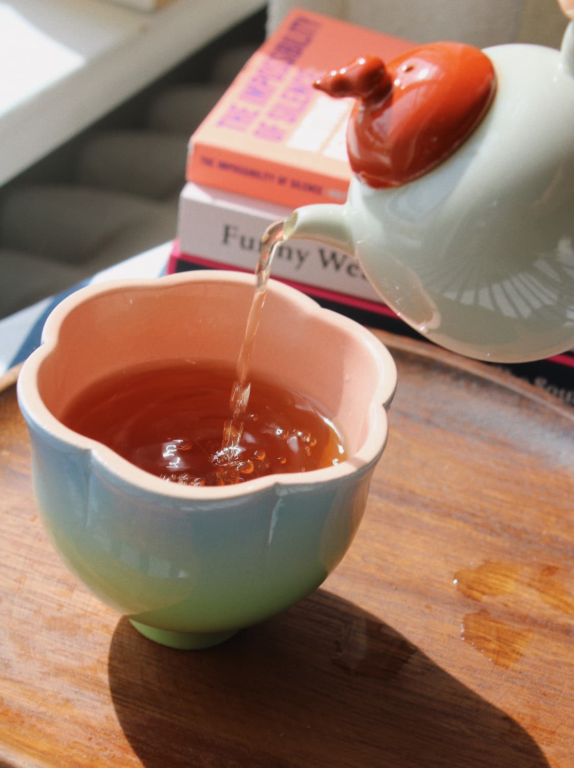 A ceramic teapot with a reddish-orange lid is pouring amber tea into a pastel gradient flower-shaped cup on a wooden tray, with stacked books blurred in the background.