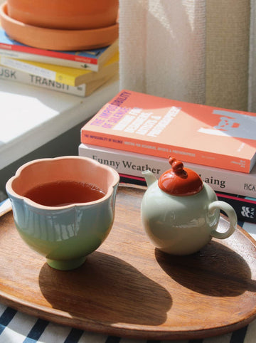A ceramic tea set with a pale green body and a terracotta-colored lid, alongside a matching flower-shaped cup filled with tea, arranged on a wooden tray next to stacked books in a sunlit indoor setting.
