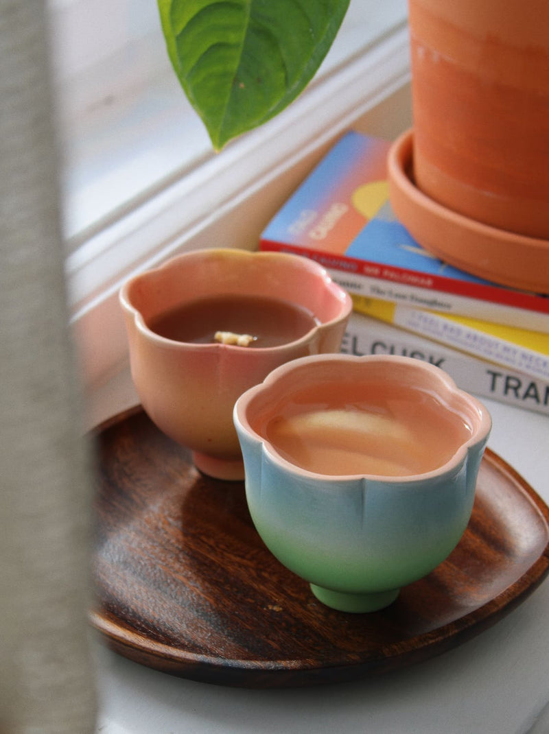 Two small ceramic cups with scalloped flower-shaped rims sit on a dark wooden tray on a windowsill. One cup is peachy pink and the other is blue-green to green, both containing a light brown liquid.