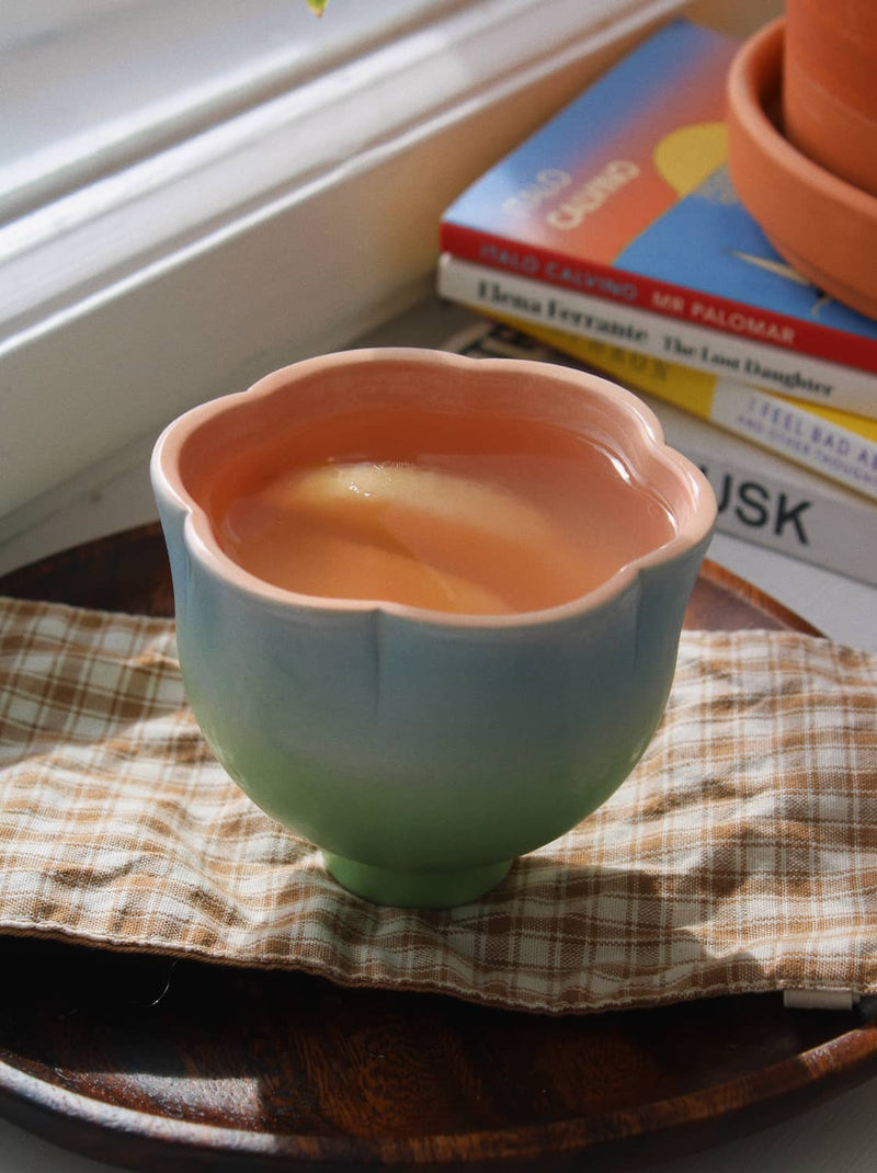 A ceramic cup with a scalloped rim, featuring a color gradient from peach-pink at the top to light blue and mint green at the bottom, resting on a plaid napkin on a wooden tray.