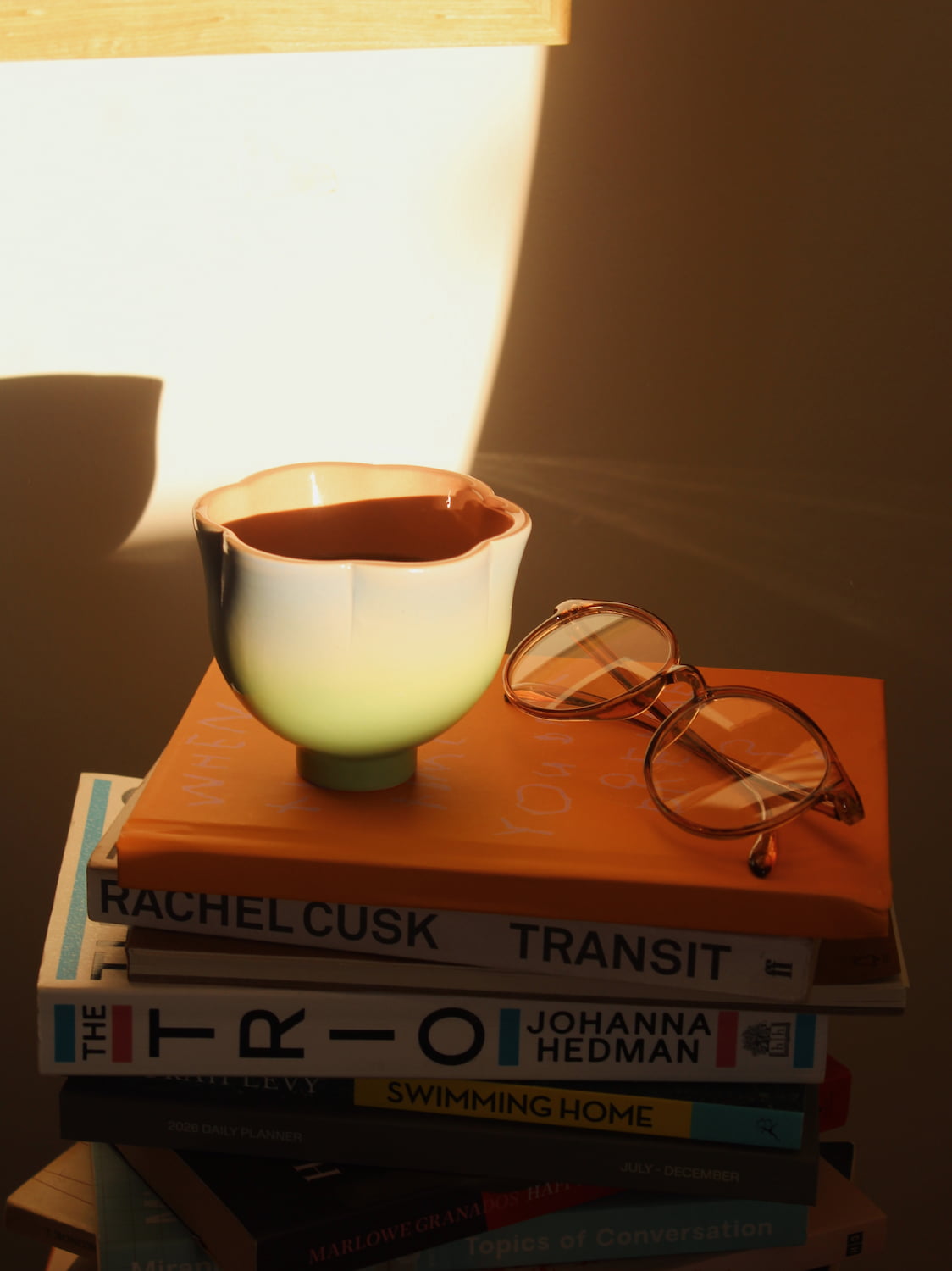 A small ceramic bowl with a white and pale green body and a terracotta-colored interior sits on top of a stack of books, including Rachel Cusk Transit and The 1800, with a pair of round eyeglasses resting beside it in warm, dramatic lighting.