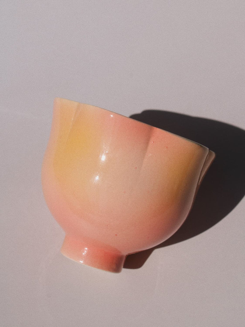 A glossy ceramic bowl with a soft peach-pink and pale yellow gradient glaze, photographed on a light gray background with a sharp cast shadow.