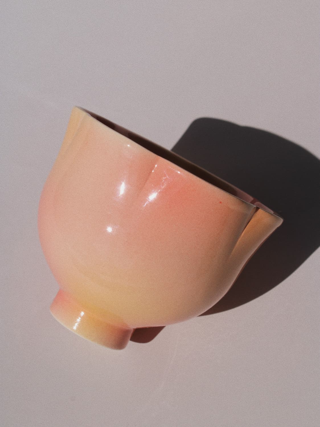 A glossy ceramic bowl with a soft peach-pink to pale yellow gradient glaze, photographed on a light neutral background with a strong cast shadow.