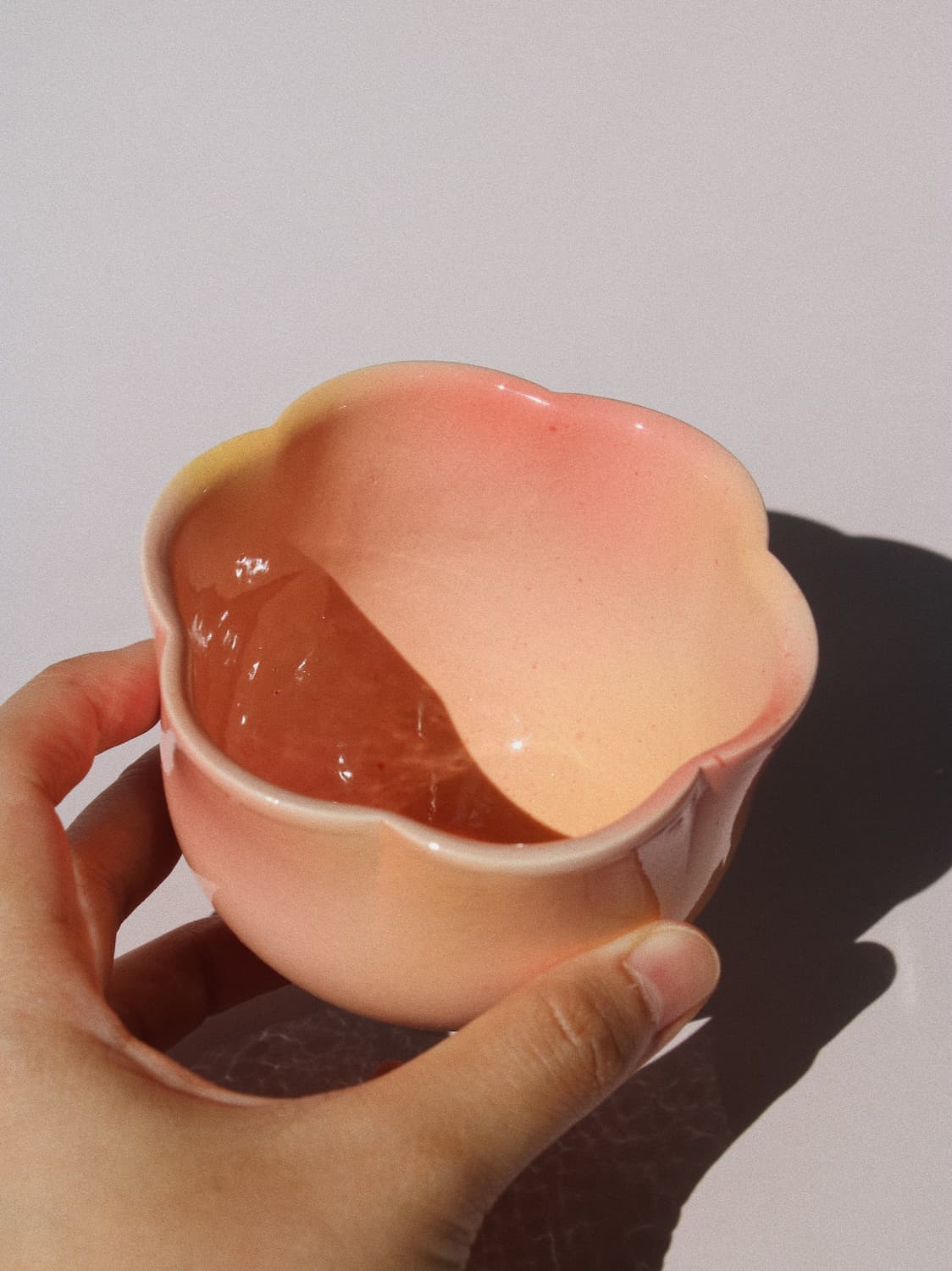 A hand holding a small flower-shaped ceramic cup with a pink and peach gradient glaze, casting a shadow on a light gray background.
