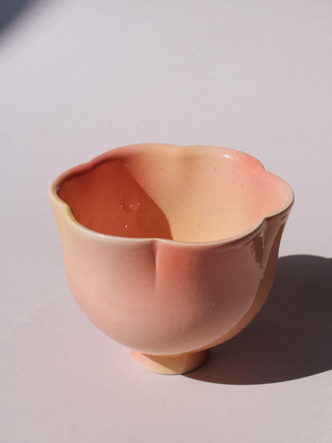 A small ceramic cup with a scalloped rim and a short pedestal base, shown on a plain light background. The cup has a glossy finish with a color gradient shifting from a warm peach-pink on the exterior to a deeper terracotta-orange on the interior.