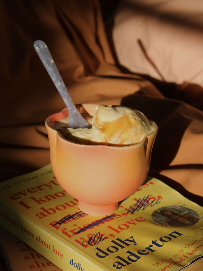 A peach-colored ceramic bowl filled with vanilla ice cream and topped with caramel drizzle, with a translucent plastic spoon resting inside. The bowl is placed on top of a yellow book titled 'dolly alderton' against a background of draped brown fabric.