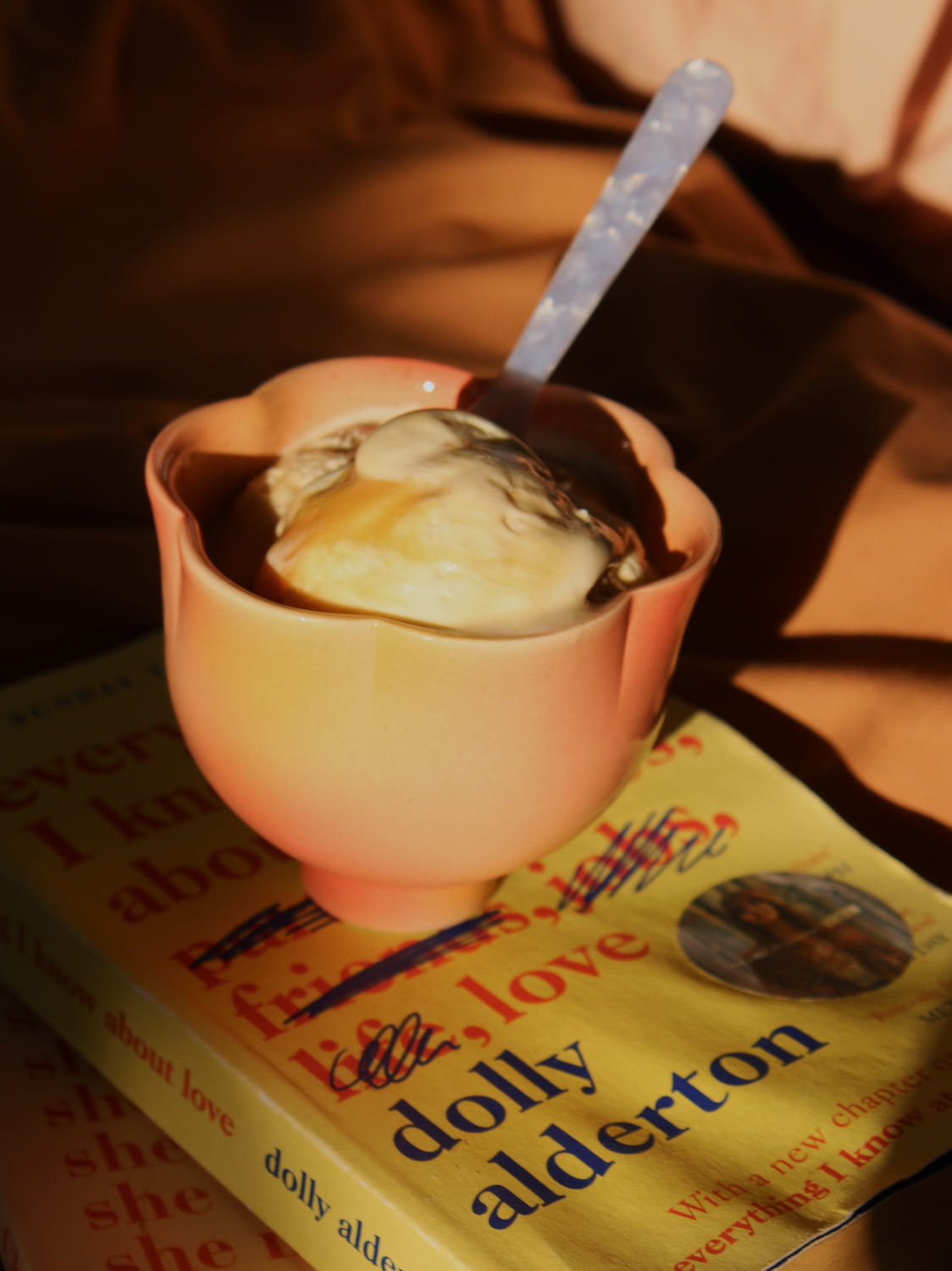 A small peach-colored ceramic bowl filled with ice cream and topped with a spoon, resting on top of a yellow book titled 'dolly alderton' in warm, low-light lighting.
