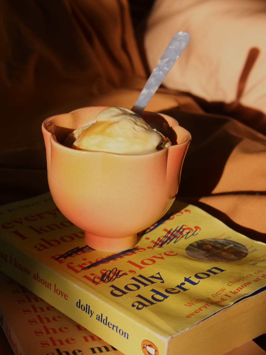 A peach-colored ceramic cup filled with vanilla ice cream and a blue-handled spoon, resting on top of two yellow books titled 'I Know About Love' by Dolly Alderton in warm, dim lighting.