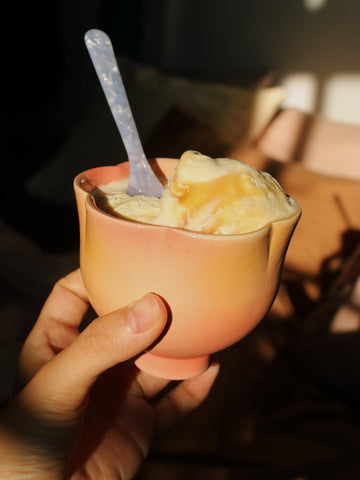 A hand holding a small peach-colored ceramic cup filled with vanilla ice cream and topped with caramel drizzle, with a translucent blue spoon inside. The background is dark and softly blurred with warm light patches.