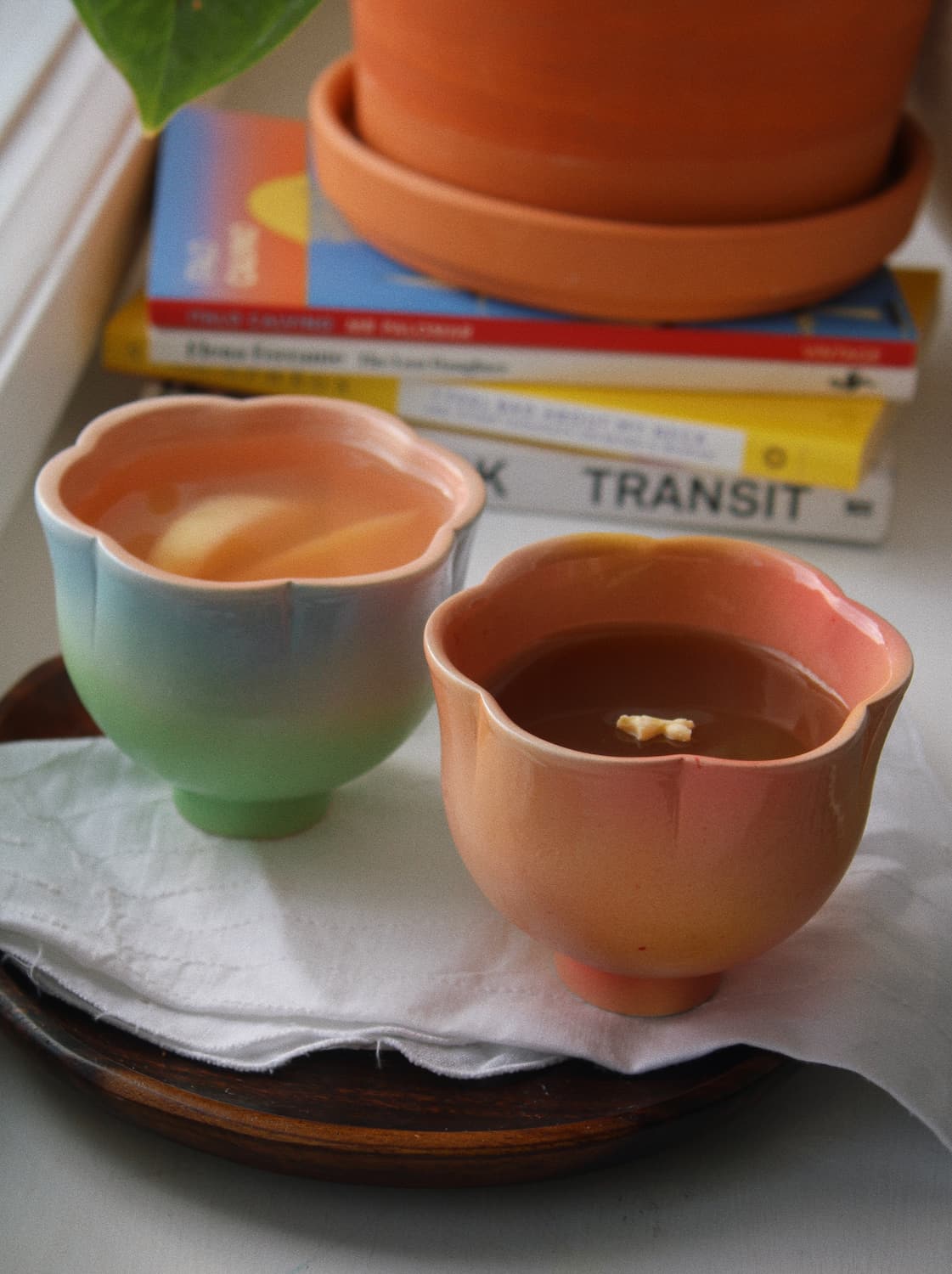 Two handmade ceramic cups with scalloped edges, one showing a blue-to-green gradient and the other a peach-to-orange gradient, filled with tea and placed on a white napkin on a wooden tray.