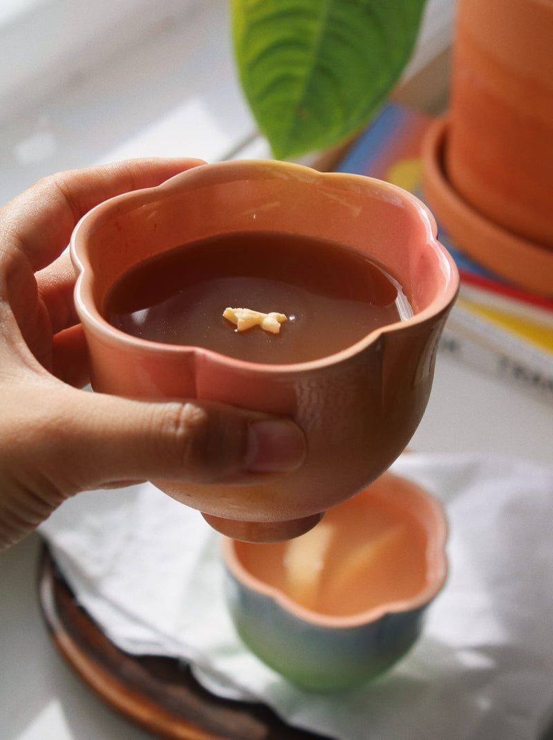 A hand holds a small scalloped ceramic cup filled with a dark brown beverage, with another matching cup blurred in the background on a white cloth.