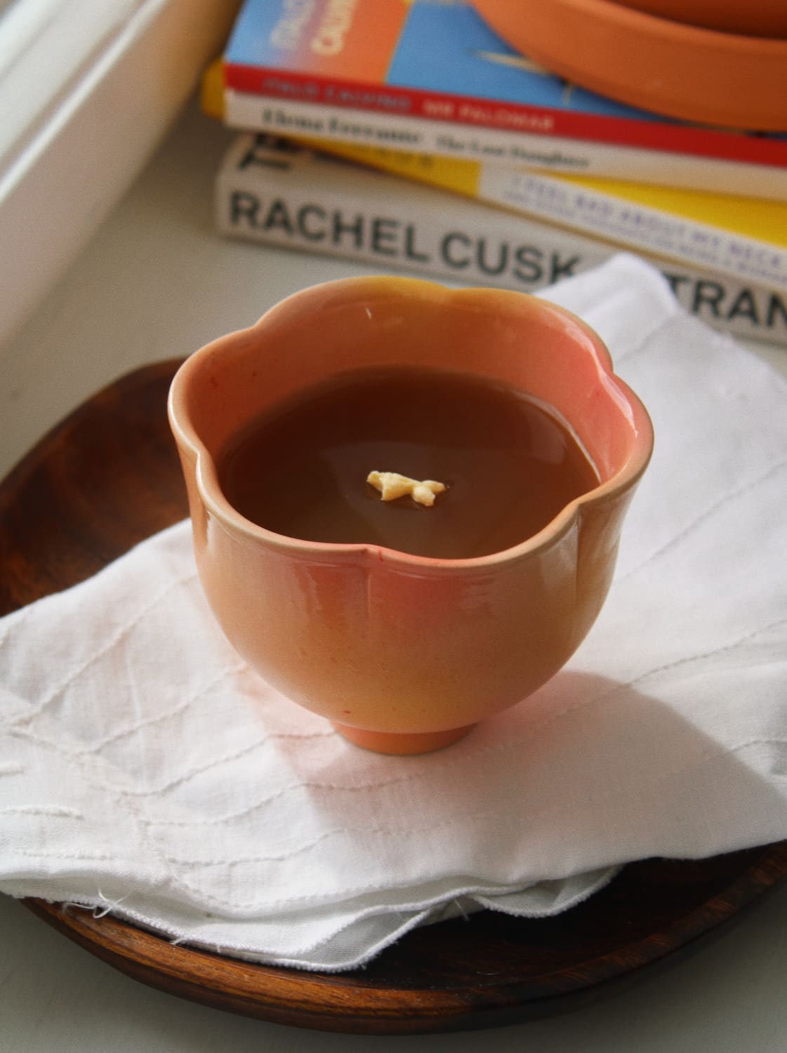 A small scalloped ceramic bowl filled with dark brown soup, resting on a white cloth over a wooden plate, with books in the background.