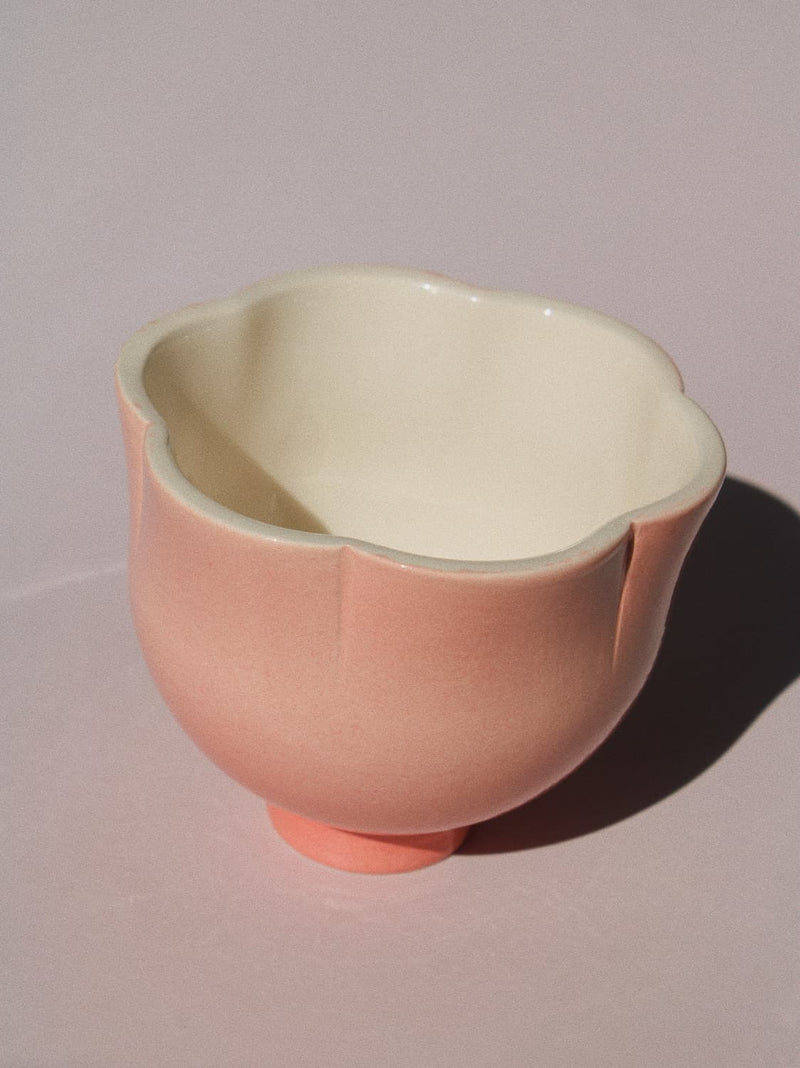 A small ceramic cup with a scalloped rim and a short pedestal base, shown on a plain light background. The exterior is a soft blush pink, and the interior is a warm cream color.