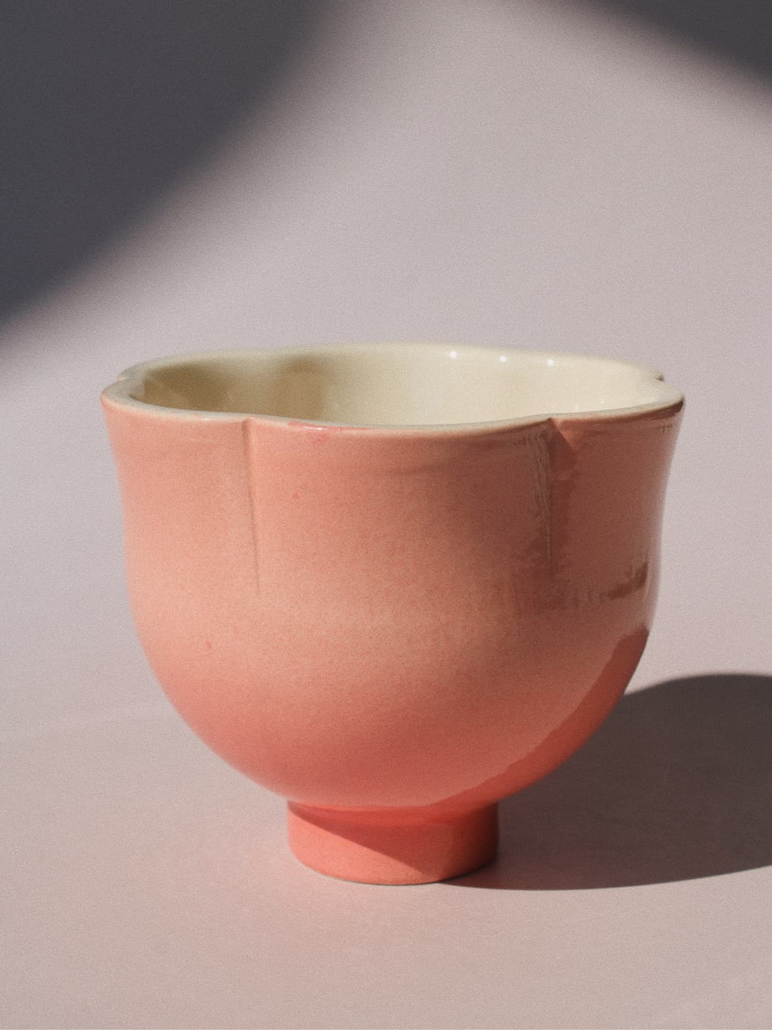 A handmade ceramic cup with a scalloped rim and a small pedestal base, featuring a soft pink exterior glaze and a contrasting cream-colored interior, set against a neutral light gray background with artistic shadows.