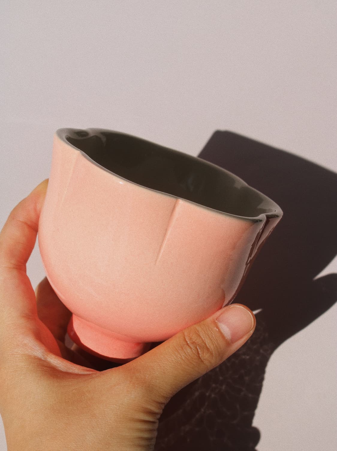 A hand holding a small glossy ceramic cup or bowl in a soft blush pink color, with a dark interior, against a plain light background with a strong shadow.
