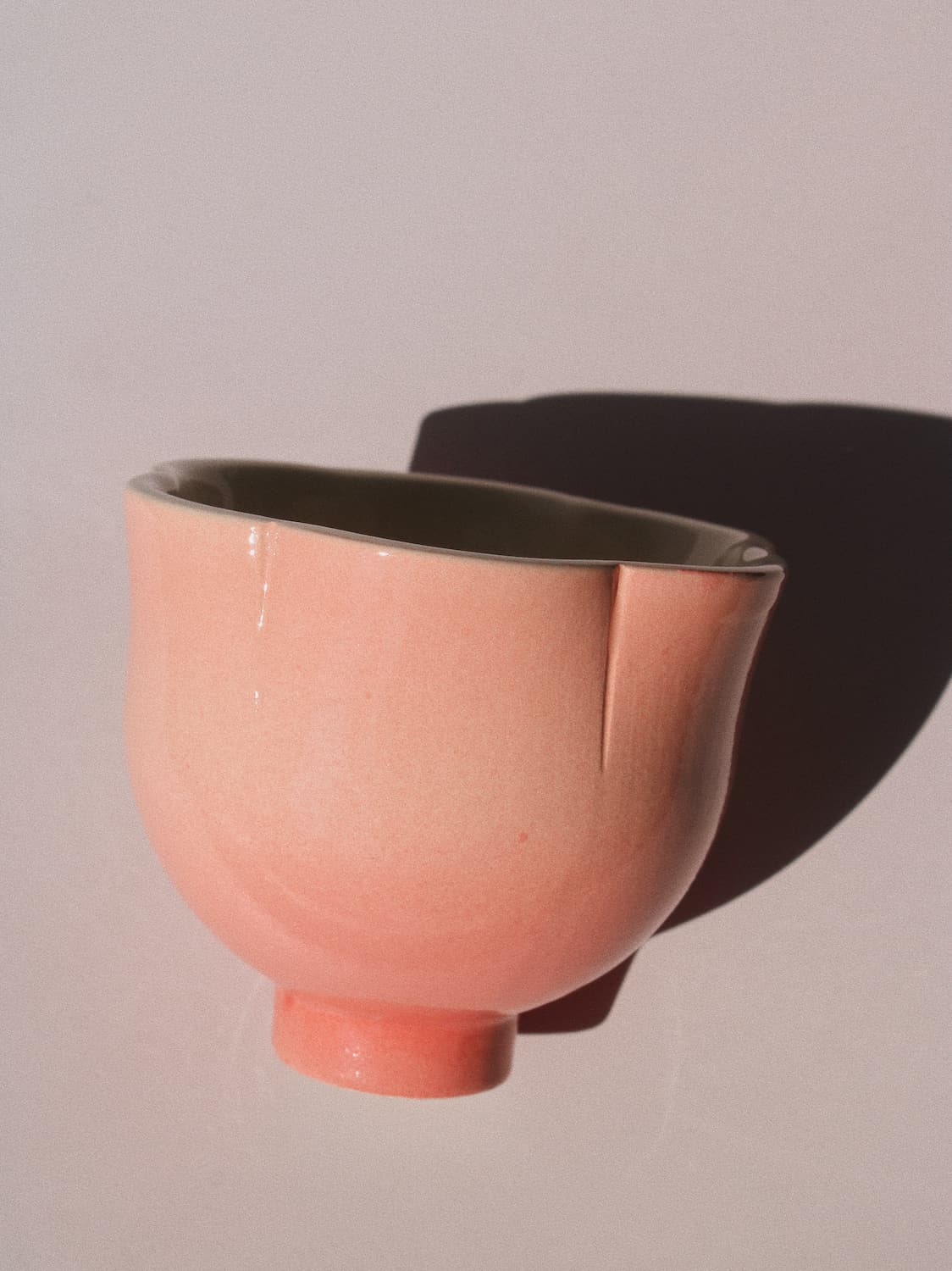 A glossy ceramic bowl in a soft peach-pink color with a dark interior, photographed against a light neutral background with a prominent shadow.