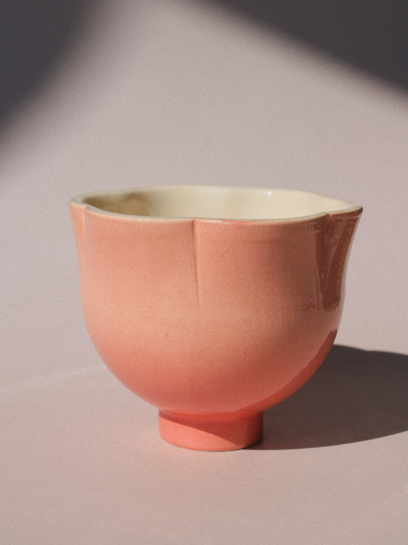 A ceramic cup with a fluted rim and a small pedestal base, shown in a soft peach-pink exterior with a warm cream interior, photographed on a neutral surface with directional sunlight and shadow.