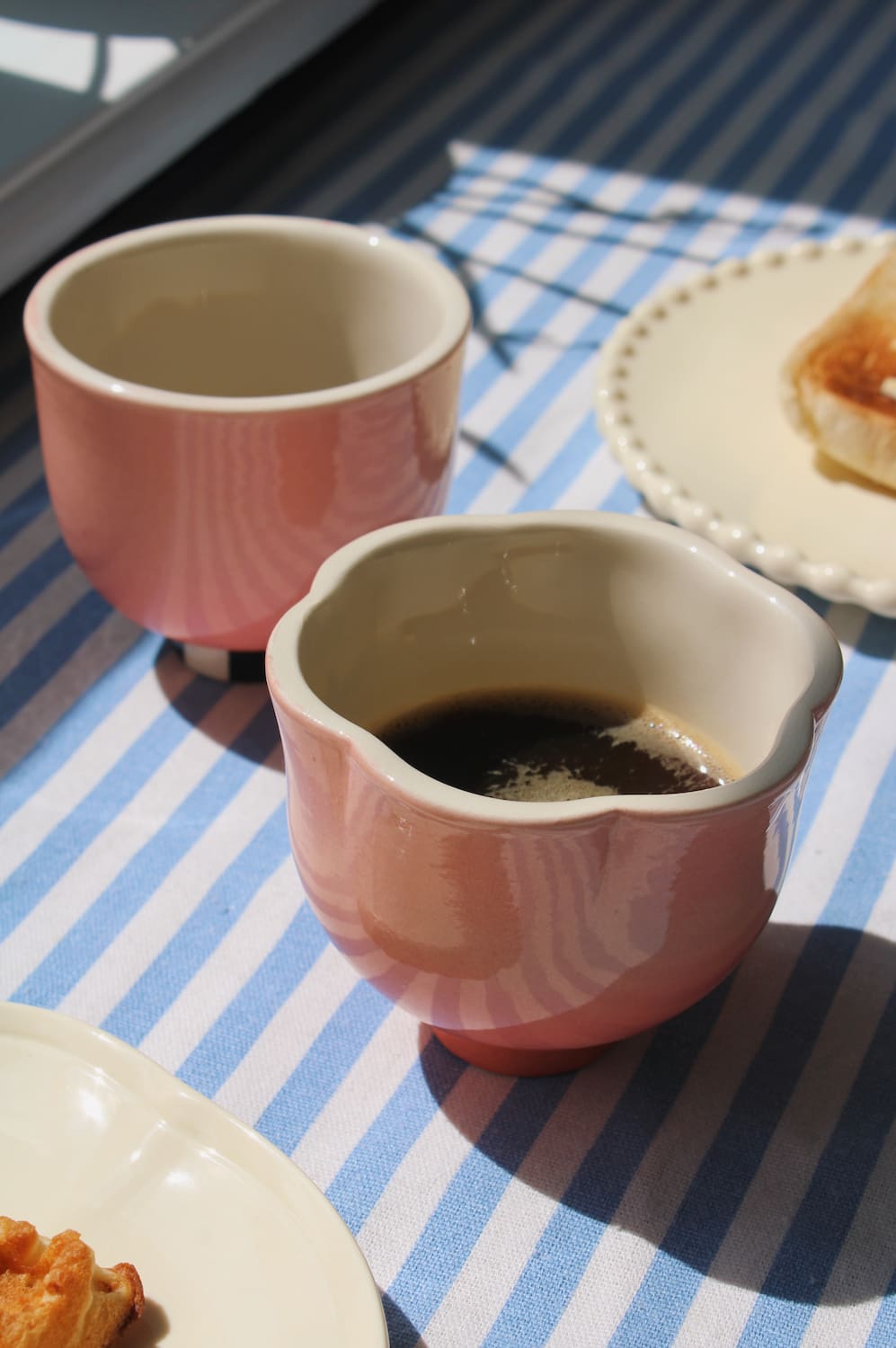 Two pink ceramic cups with scalloped edges, one filled with dark coffee and the other empty, set on a blue and white striped tablecloth with plates of food nearby.