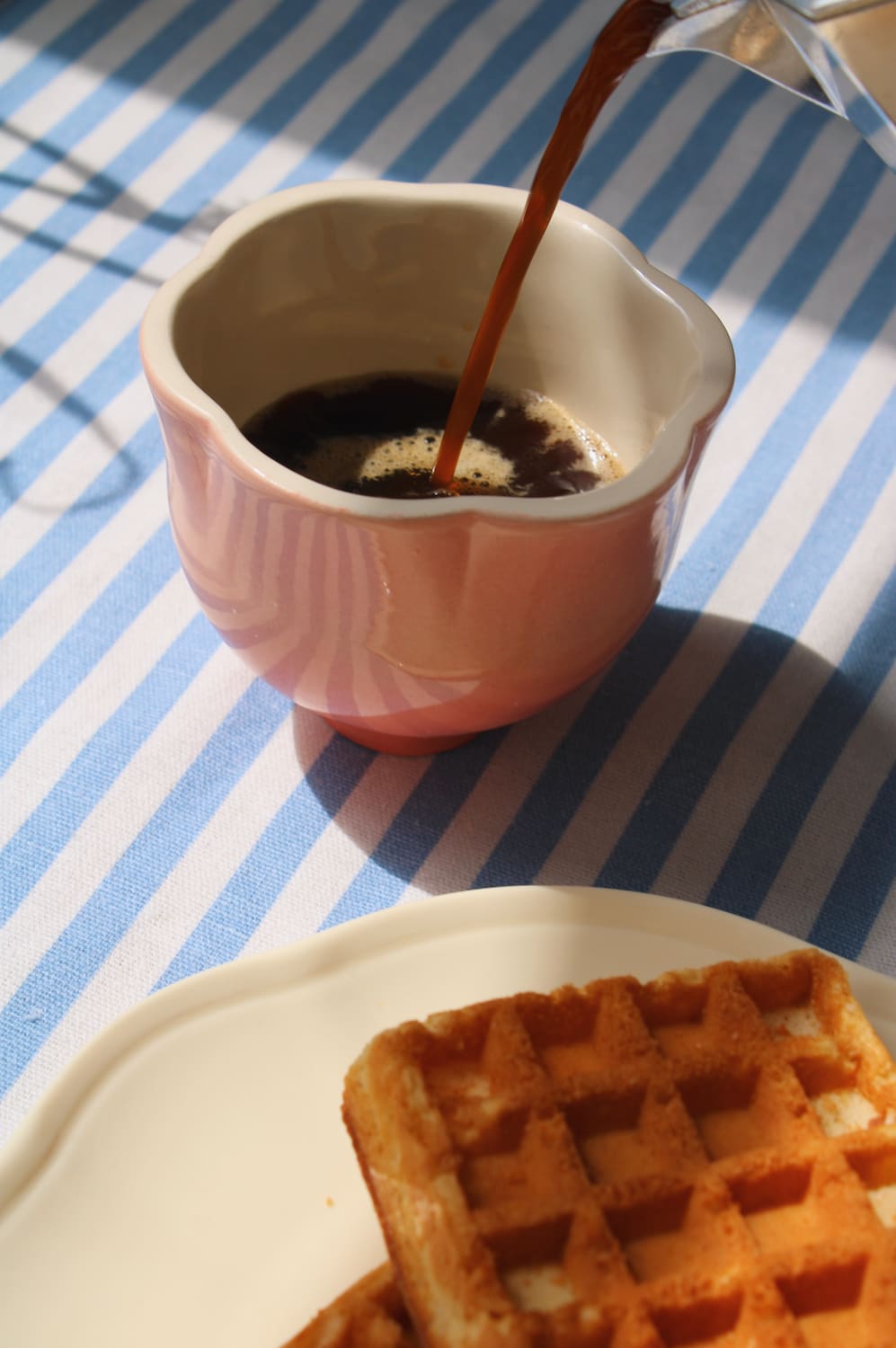 A pink ceramic pitcher filled with coffee, with coffee being poured into it, set on a blue and white striped tablecloth next to a plate of waffles.