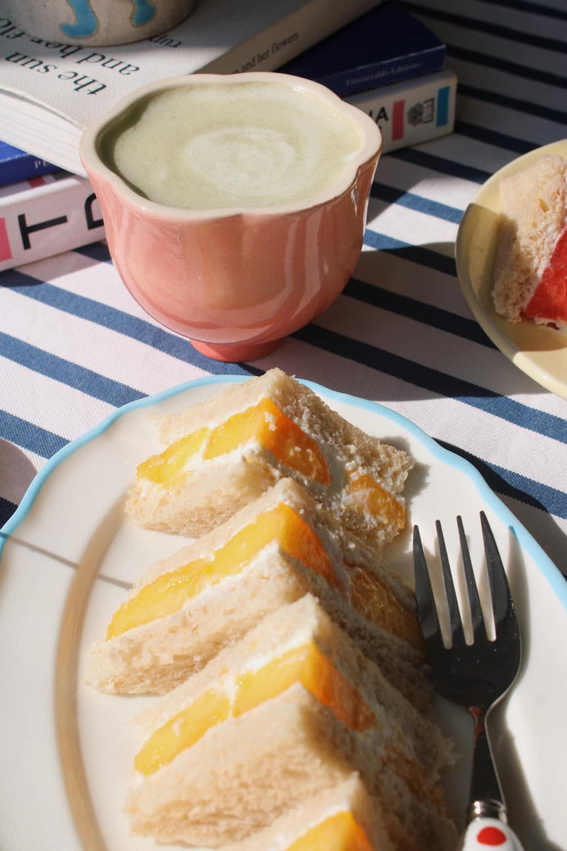 A close-up of a pink ceramic cup filled with a light-colored beverage, alongside a plate of sandwich cake slices with yellow fruit filling and a fork, set on a striped tablecloth with books in the background.