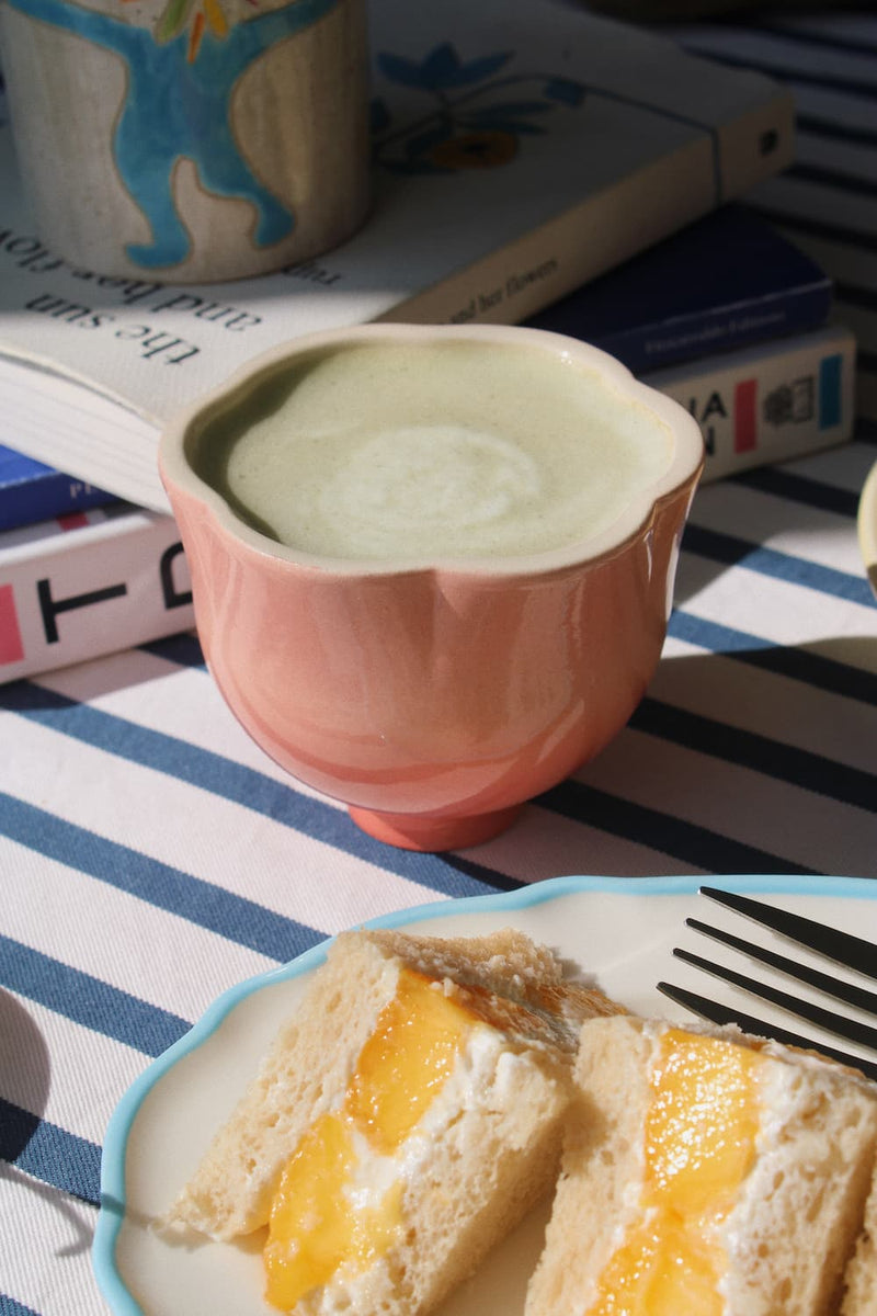 A pink ceramic cup filled with a pale green drink sits on a striped tablecloth beside a plate with sandwich bread and yellow fruit filling, with books and a mug in the background.