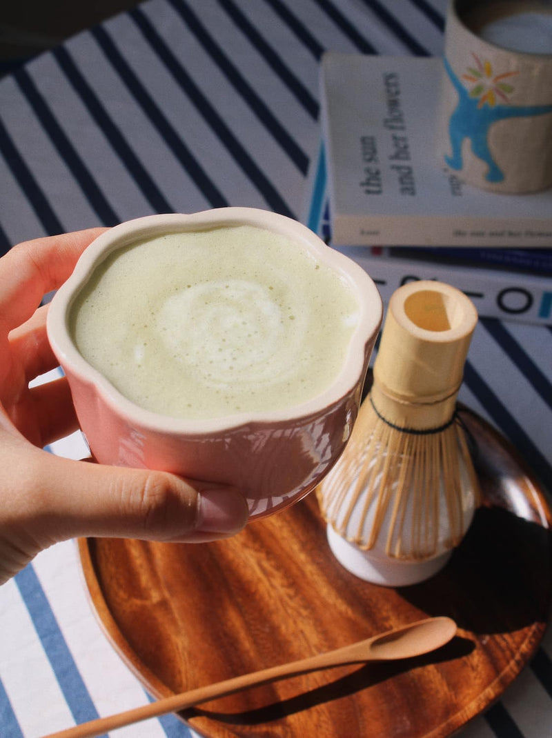 A hand holds a pink ceramic cup filled with a light green matcha drink, set on a wooden tray beside a bamboo whisk and spoon, with a striped cloth and books in the background.
