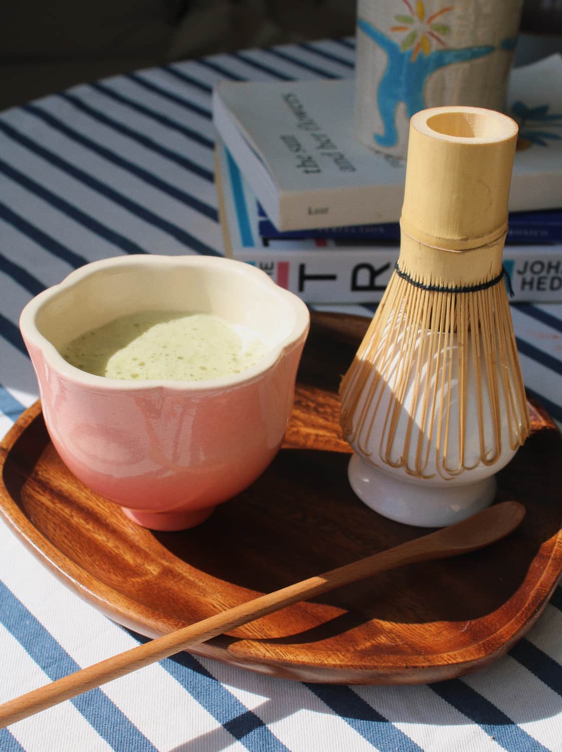 A pink ceramic matcha bowl filled with green tea sits on a wooden tray beside a bamboo whisk, with a wooden spoon and stacked books in the background on a blue-and-white striped tablecloth.