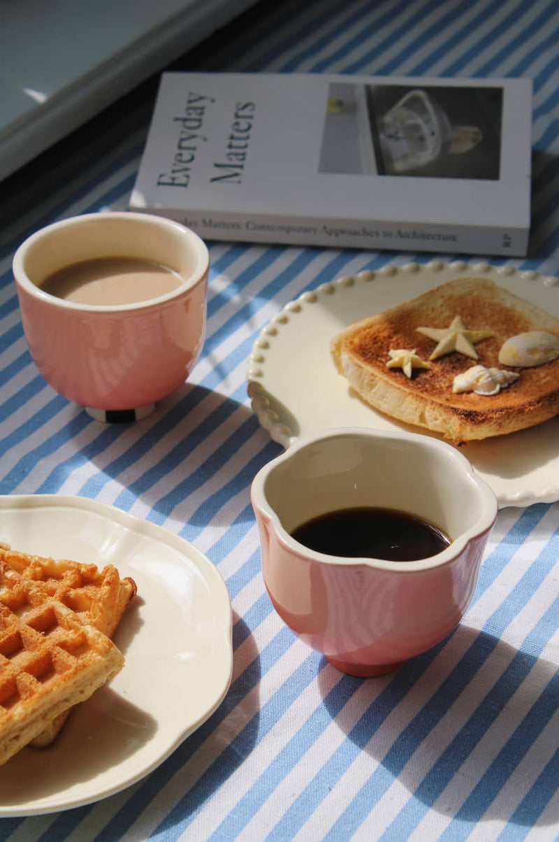 A cozy breakfast scene with two pink ceramic cups, one filled with coffee and the other with a light brown drink, set on a blue-and-white striped tablecloth alongside plates of waffles and toast topped with star-shaped cookies. A book titled 'Everyday Matters' lies in the background.