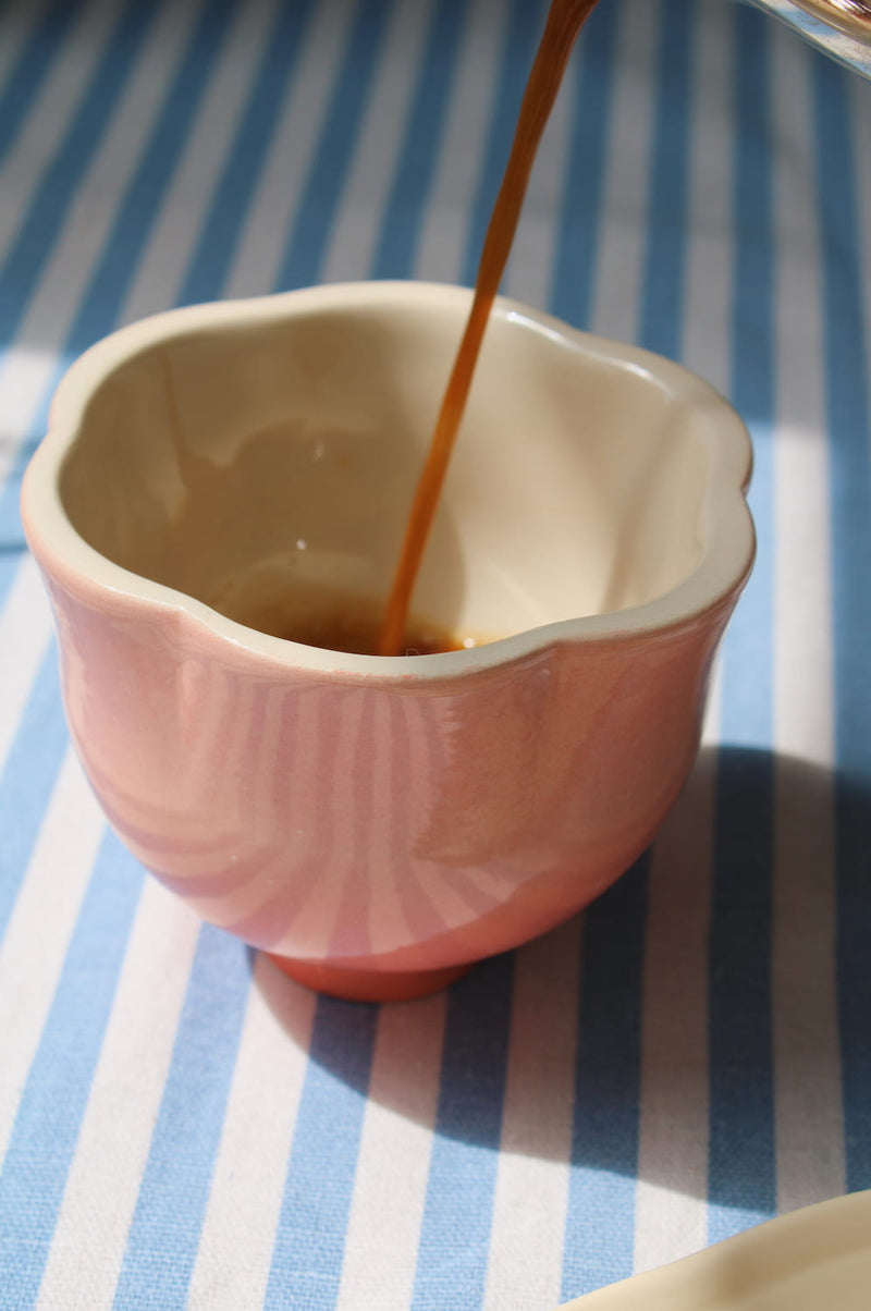 A pink ceramic cup with a scalloped rim and ribbed texture, shown on a blue and white striped surface with a dark liquid being poured into it.