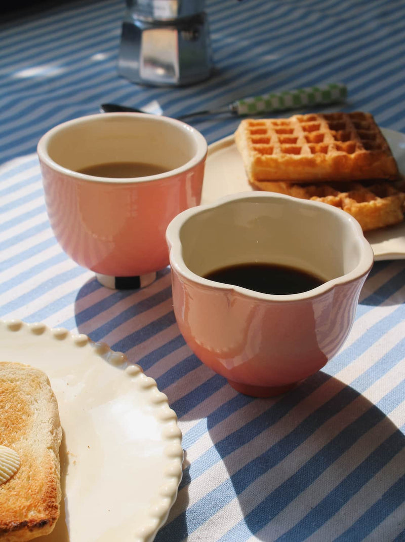 Two pink ceramic cups filled with coffee on a blue and white striped tablecloth, with waffles and toast in the background.