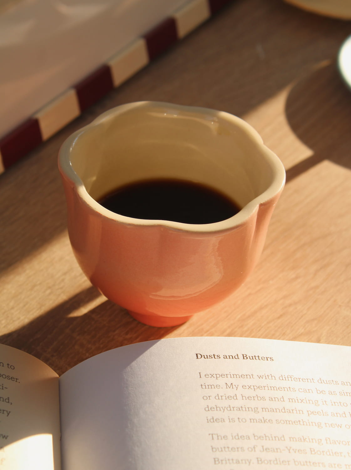 A small ceramic cup filled with black coffee sits on a wooden table next to an open book, with warm sunlight casting shadows.