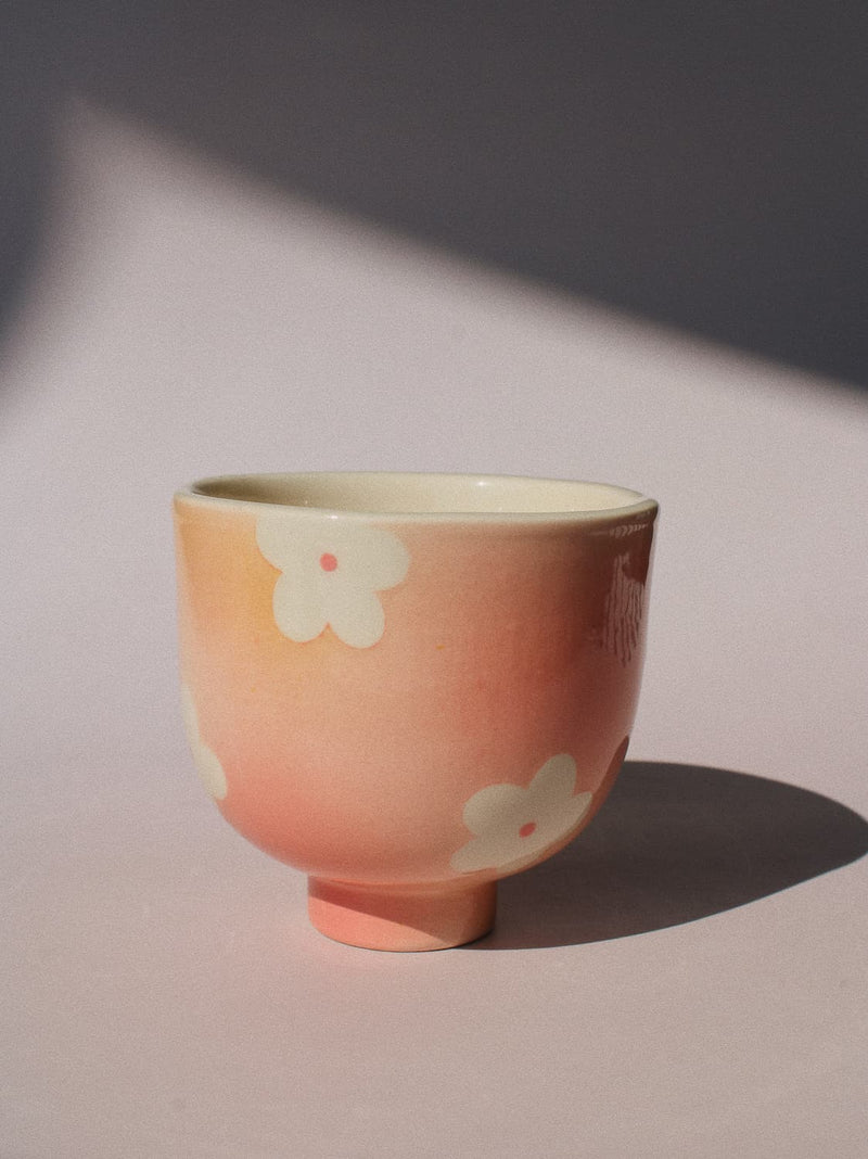 A small ceramic cup with a pink and peach gradient, decorated with white cherry blossom flowers, set against a neutral background with soft shadows.