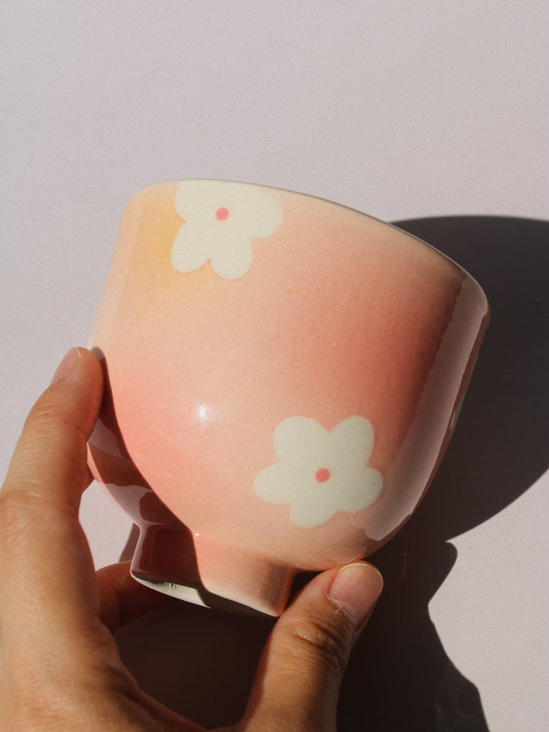 A hand holding a glossy pink ceramic cup decorated with white flower motifs, set against a plain light background with a strong shadow.