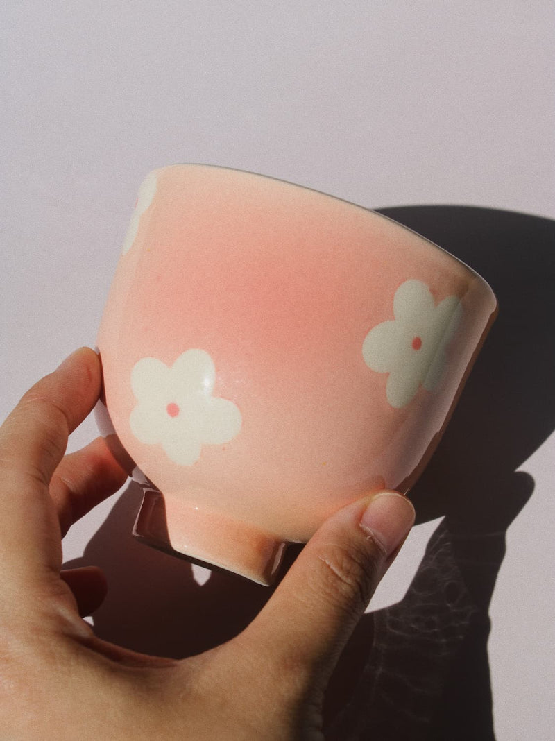 A hand holding a glossy pink ceramic bowl decorated with white cherry blossom flowers, set against a plain light background with a strong shadow.