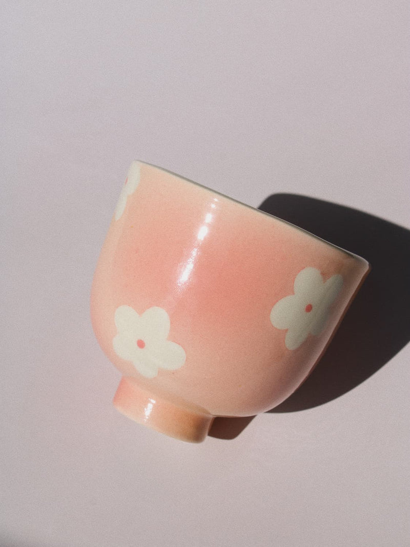 A light pink ceramic cup decorated with white hand-painted cherry blossoms, shown on a neutral light gray background with a soft shadow.