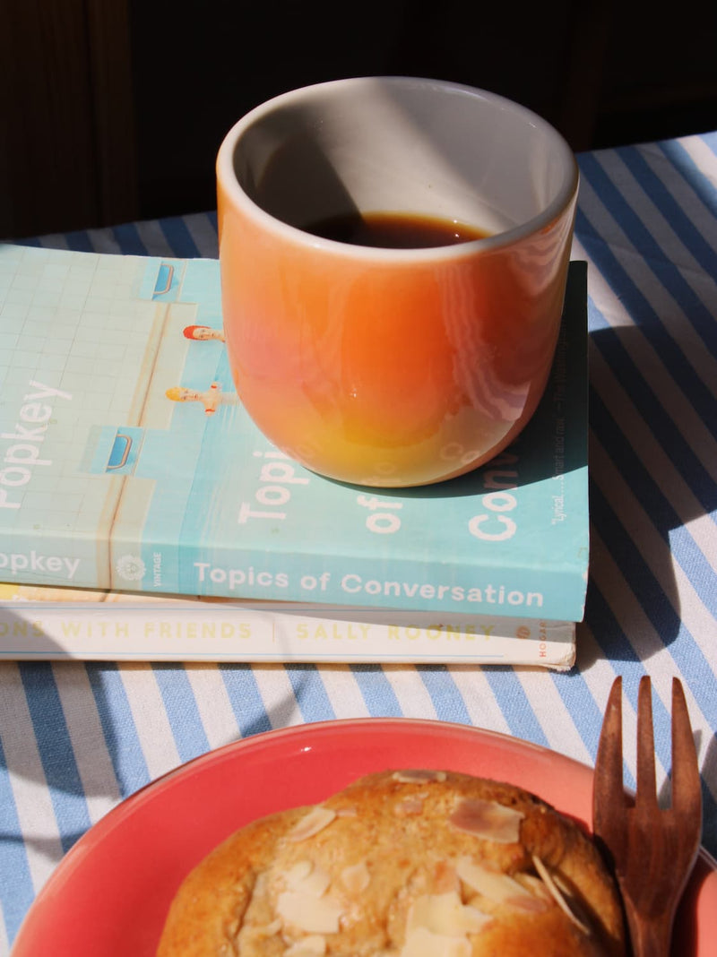 Sunset Gradient Ceramic Cup by Agata Puławska – colourful handmade ceramic cups styled with coffee and a pastry on a blue striped tablecloth