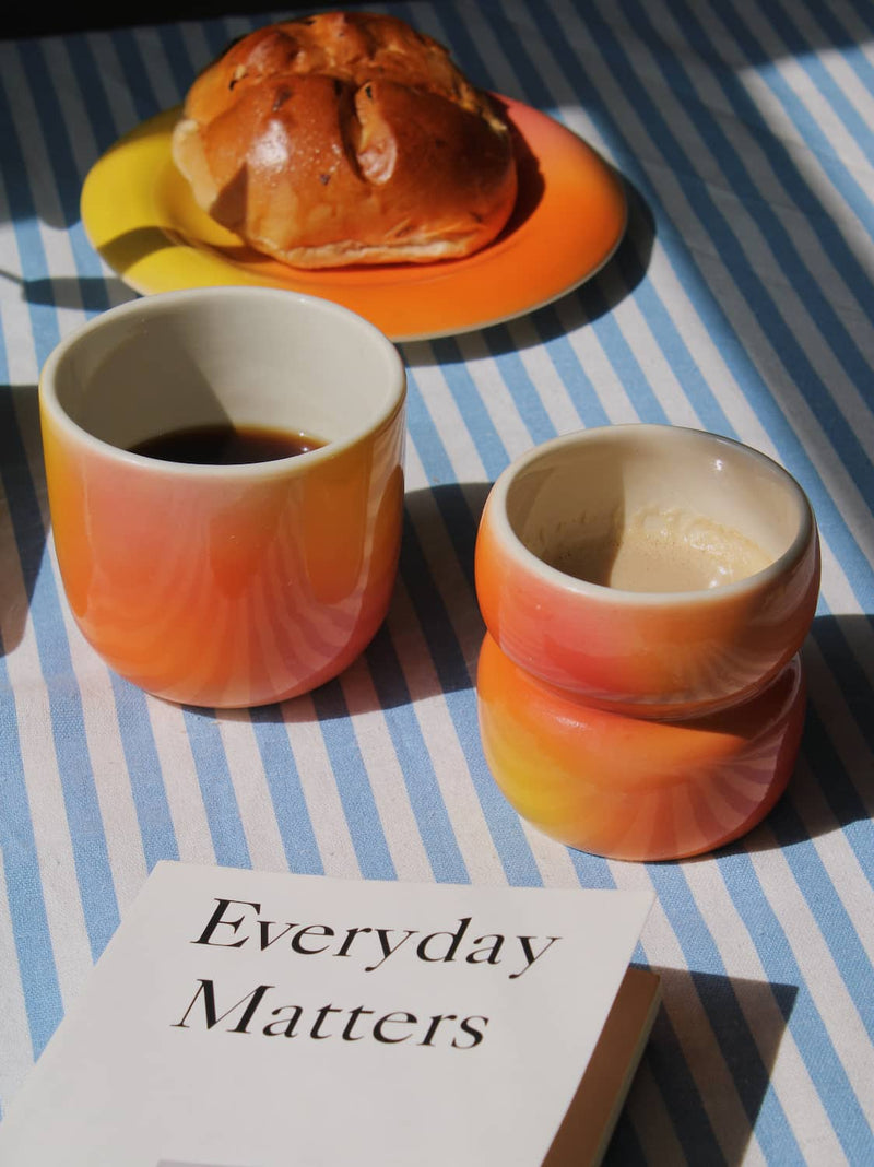 Sunset Gradient Ceramic Cup by Agata Puławska – colourful handmade ceramic cups styled with coffee and a pastry on a blue striped tablecloth