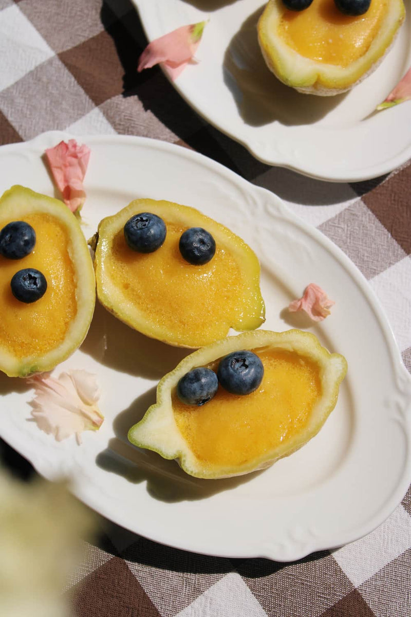 Auriel oval serving plate in white ceramic with scalloped rim, styled with mango halves and blueberries on a checkered linen