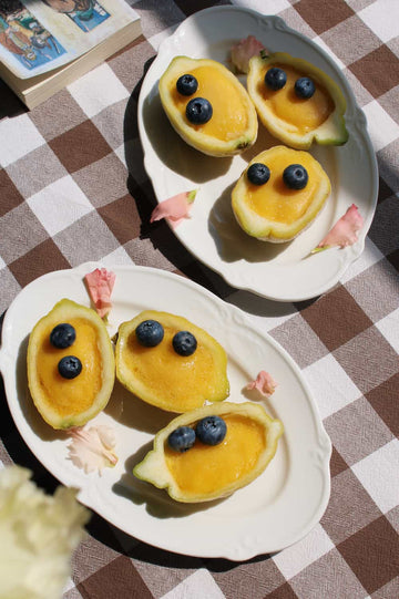 Auriel oval serving plate in white ceramic with scalloped rim, styled with mango halves and blueberries on a checkered linen