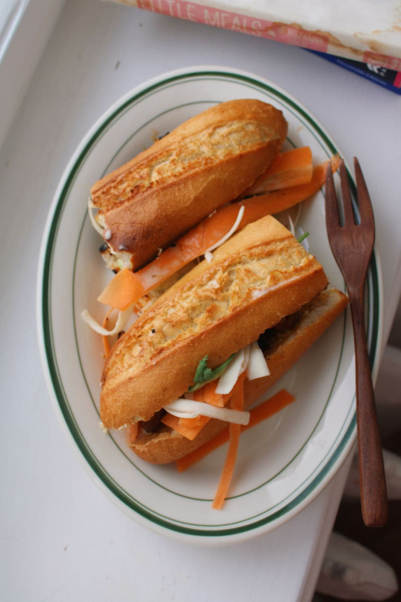 Two baguette-style sandwiches filled with shredded cabbage and carrot slices, served on a white oval plate with green trim and a wooden fork, set on a white surface.