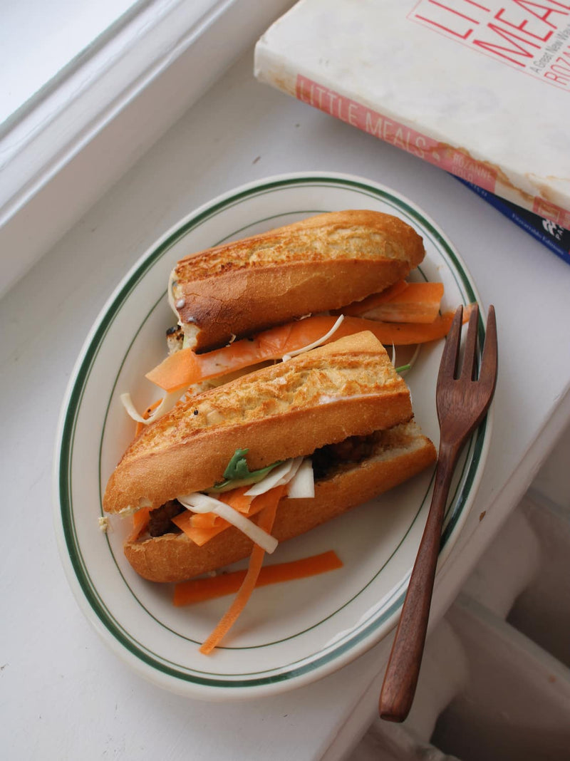 Two baguette-style sandwiches filled with shredded cabbage and carrot slices, served on a white oval plate with green trim and a wooden fork, set on a white surface.