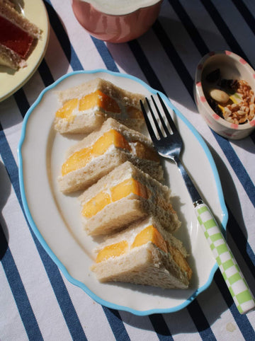 A plate of four triangular sandwich slices filled with orange fruit, served on a white oval plate with a light blue rim and a green-and-white checkered fork handle, set on a blue-and-white striped tablecloth.