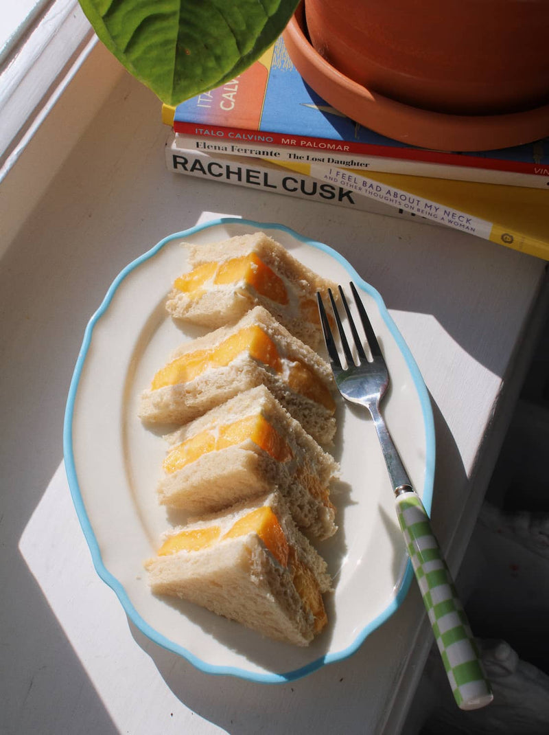 A plate of four triangular sandwich slices with orange-yellow filling, served on a white oval plate with a light blue rim and a green-and-white checkered fork handle, set on a windowsill with sunlight and books in the background.