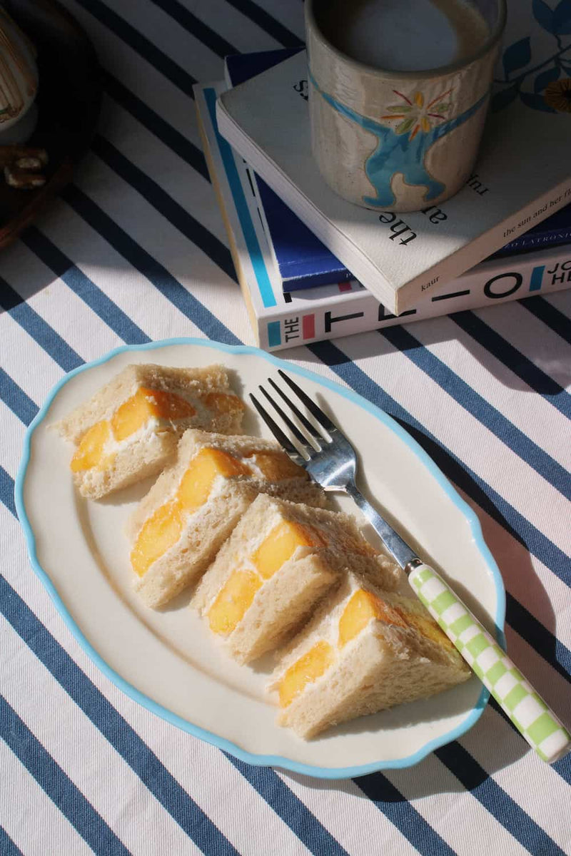 Four triangular slices of sandwich bread with yellow fruit filling, served on a white scalloped plate with a fork, set on a blue-and-white striped tablecloth next to stacked books and a ceramic mug.