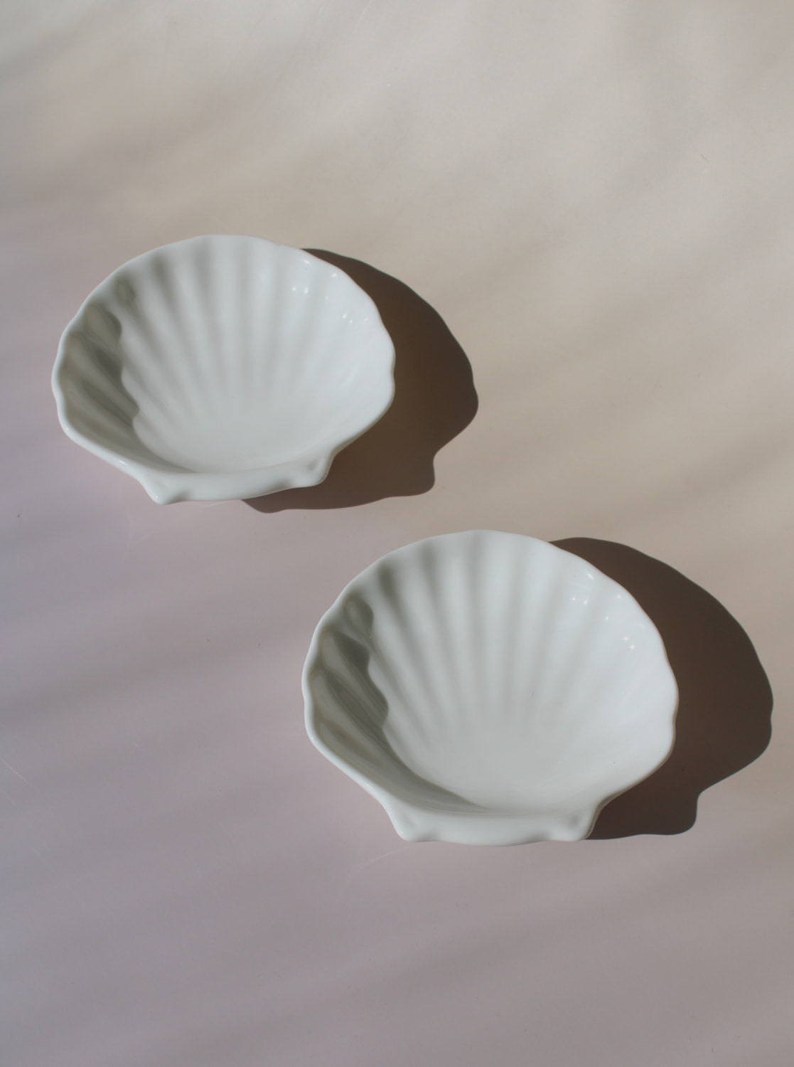 Two white ceramic shell-shaped bowls on a light pink background