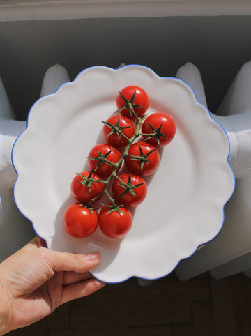 White scalloped-edge plate with red tomatoes held by a hand against a neutral background